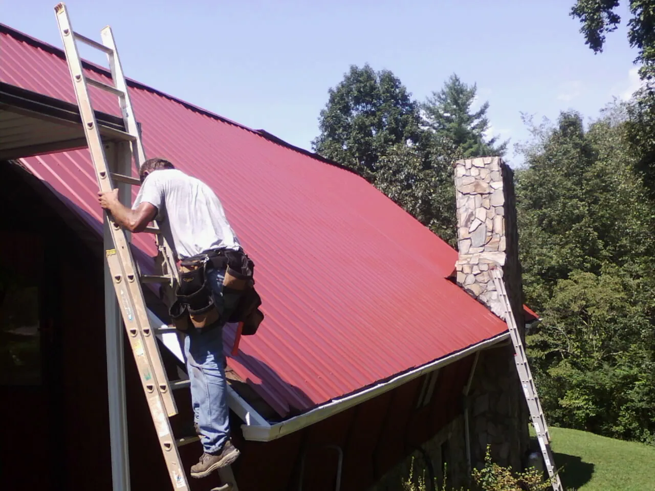 Roofer on a ladder beside a red metal roof and stone chimney — Soffit & Fascia Repair by Roofing Brothers in Half Moon Bay